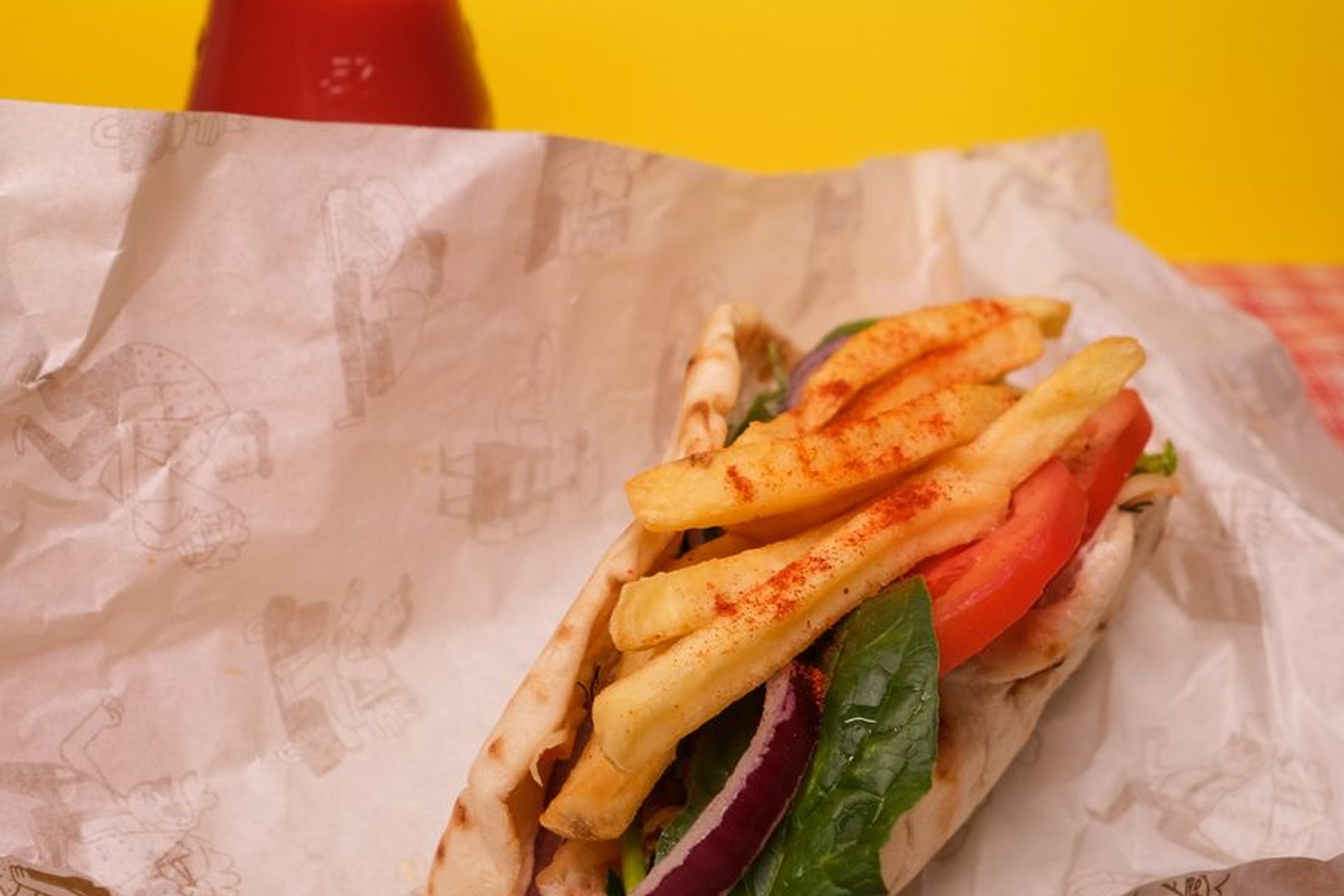 Sárga napszemüveges, színes kabátos fiatal nő beleharap egy gyros pitába egy graffitis fal előtt.