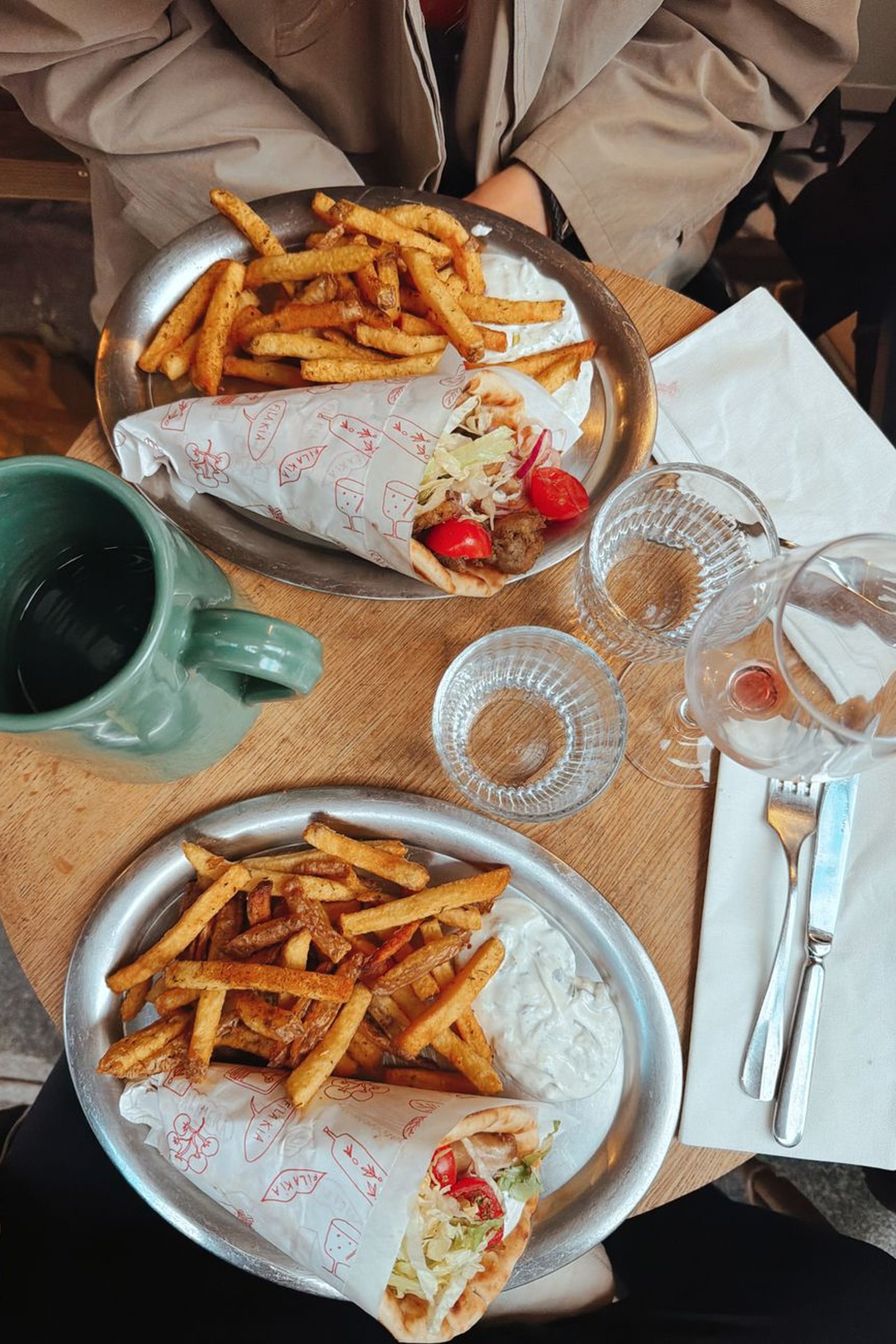 Vidám, szőke fiatal nő két gyros tálat tart az arca mellé és szélesen mosolyog egy beltéri fal előtt.